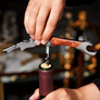 A close-up of a person using a corkscrew opener with a wooden handle and silver metal wings to remove a cork from a wine bottle, with a blurred bar or kitchen background.