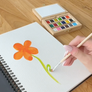 A hand holds a paintbrush, painting a green stem on an open white spiral sketchbook. An orange flower is already painted on the page.