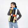 A woman modeling a chunky knit scarf with a bold geometric patchwork pattern in black, yellow, blue, orange, and white, styled over a dark gray long-sleeve shirt and light blue jeans against a plain white background.