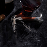 A corkscrew wine opener with a polished silver metal body and a brown wooden handle, resting on a dark, textured surface with wine glass props in the background.