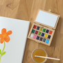 A wooden watercolor paint palette with multiple colored pans, an open blank white canvas, and a small cup of yellow paint with a brush, arranged on a light wood floor.