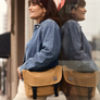 Woman in a denim jacket and white pants stands against a window, wearing a camel Classic Messenger Bag with black straps. Her reflection is visible in the glass behind her.