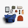 A styled flat-lay of multiple accessories arranged on a white background next to indigo Classic Messenger Bag.