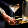 A Deco Gold Rimmed Martini Glass with an olive garnish, held by a hand on a gold tray next to a ribbed clear glass bottle against a dark background.