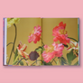An open art book laid flat against a solid pink background, featuring a floral cover design with pink and red flowers on a mustard yellow background.