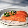Two salmon fillets on a white plate with a sprig of green parsley and scattered peppercorns, photographed in a bright, softly blurred setting.