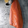 A large raw salmon fillet laid on a gray stone surface, with lemon halves, a small jar of salt, and scattered coarse salt and black pepper nearby.