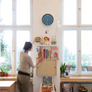 A woman stands in a bright room beside large windows, reaching toward a wall-mounted light wood bookshelf filled with books and small decor items. A blue round wall clock hangs above the shelf, and the room has a warm, sunlit interior with plants and wooden surfaces.