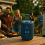 A dark blue portable Bluetooth speaker with a carrying loop sits on a wooden table outdoors, with a blurred backyard party scene in the background.