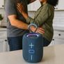 A portable blue speaker with a handle sits on a kitchen counter in front of a couple dancing in a bright kitchen.