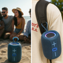 A portable blue speaker with a carrying loop is shown on a beach next to a couple, and also hanging from a person's shoulder strap. The speaker has a textured mesh front, a blue ring light, and a blue power button.