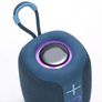 A close-up of a blue portable Bluetooth speaker with a carrying loop, featuring a purple LED ring around the speaker grille and a blue power button on the front.