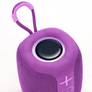 A close-up of a purple portable Bluetooth speaker with a matching purple carrying loop, featuring a black speaker grille in the center and a purple plus sign button on the side, set against a plain white background.